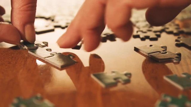 Caucasian woman hands assembling jigsaw puzzle pieces. Single pieces and complete puzzle at background. Warm colors