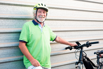 Senior people grandfather looking at camera close to his e bike. Healthy lifestyle for retired person. Yellow helmet. Metal wall in background