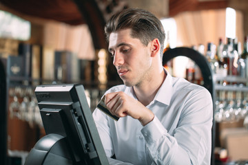 small business, people and service concept - happy man or waiter in apron at counter with cashbox working at bar or coffee shop.