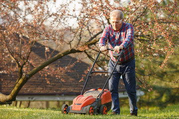 Man with electric lawnmower, lawn mowing. Gardener trimming a garden. Sunny day, suburb, village. Adult man pruning and landscaping garden, trimming grass, lawn, paths. Hard work on nature.