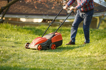 mowing trimmer - worker cutting grass in green yard at sunset. Man with electric lawnmower, lawn mowing. Gardener trimming a garden.