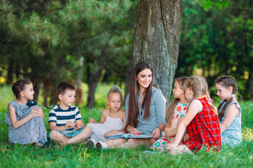 Children and education, young woman at work as educator reading book to boys and girls in park.