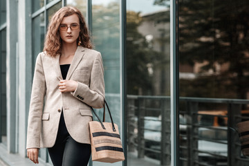 Street fashion portrait of elegant lady wearing beige blazer, oval brown  sunglasses, holding straw wicker handbag, posing in street of the city. Copy, empty space for text