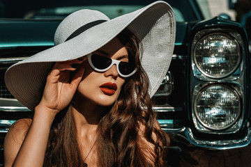 Outdoor close up portrait of young beautiful elegant lady with long luxury hair, red lips makeup, wearing trendy white cat eye sunglasses, wide brim hat, posing near retro car. Copy, empty space 