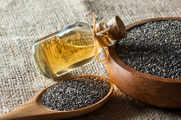 Glass bottle of chia oil and Chia seeds ( Salvia Hispanica ) in wooden spoon and bowl on burlap...