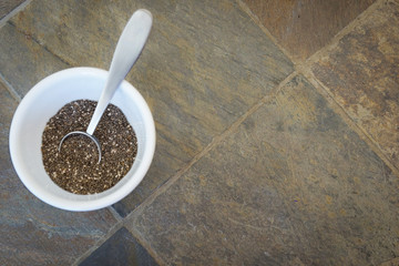 A white bowl of chia seeds with a silver metal teaspoon, on a background of brown and gray stone...