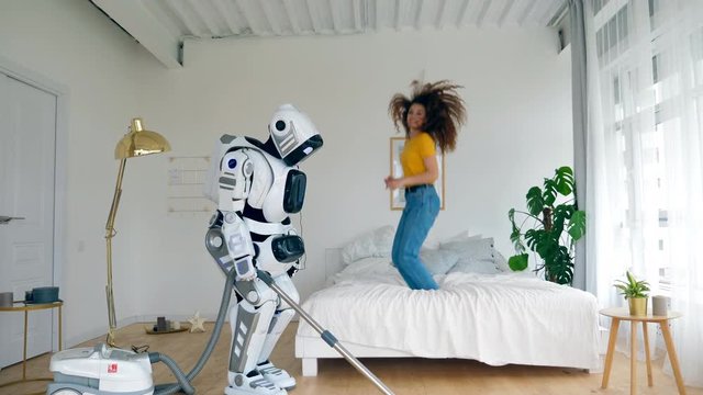 Happy girl jumps on a bed while a robot cleans floor. Robot, cyborg and human concept.