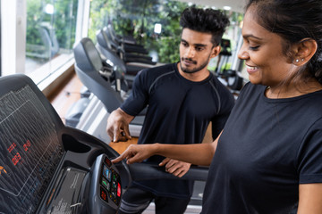Well qualified trainer explains to his client how to use treadmill, wearing black t shirt and red sports jacket. Beautiful brunette lady follows all instructions, being concentrated and attentive.