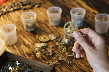 Watchmaker holds large gear. Cogwheel on blurred background of workplace. Pinion in female hand. Old cogwheel on wooden table background. Big metal gearwheel on blurred workspace.