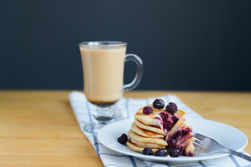 eating of fritter or oladyi with black raspberry with fork on white plate and cappuccino on table with napkin, eastern european breakfast, closeup side view of horizontal still life stock photo image