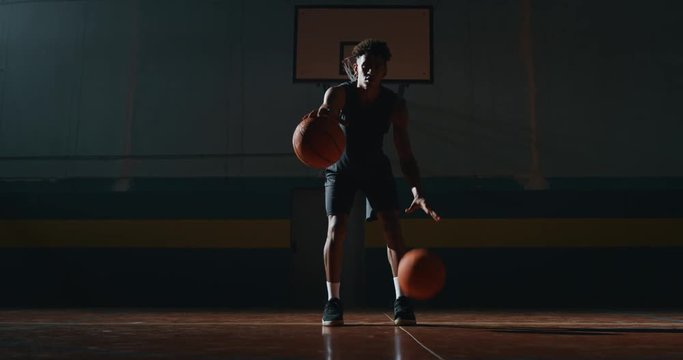 Cinematic slow motion shot of an young african professional male player is practicing a basketball work out with two balls in a gym.