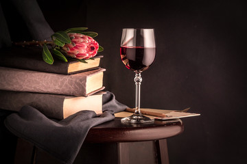 A glass of red wine, a pink protea flower and a book against a dark background. Still life with flowers. Free space for your text.