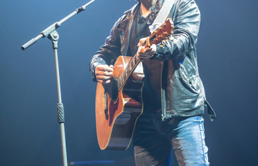 Man wearing leather jacket playing acoustic guitar on stage.