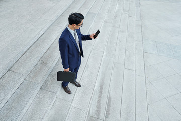 Young entrepreneur in classic suit standing outdoors and checking notifications on his smartphone, view from above
