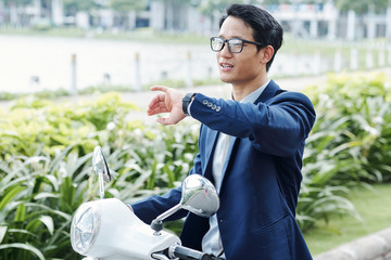 Portrait of young Asian businessman riding on scooter and checking time or notifications on his smartwatch - Powered by Adobe