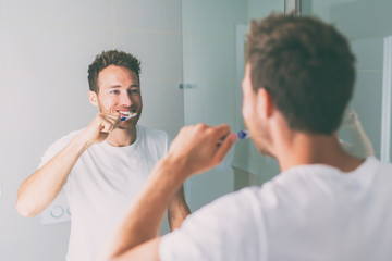 Brushing teeth man looking in mirror of home bathroom using toothbrush in morning routine for clean dental oral care.