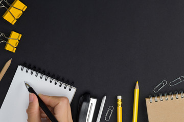 Frame from workplace with writing hand, notebook, yellow stationery on black background. Top view, flat lay, copy space, mock up