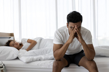 Upset depressed young man sitting on the edge of bed against his wife lying on the bed....