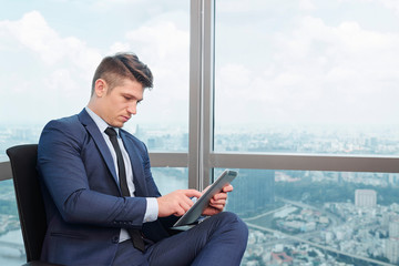 Serious young businessman sitting on chair in corner office at window with beautiful city view and working on digital tablet