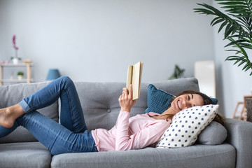 Smiling woman enjoying reading a book is lying on the sofa in the living room.