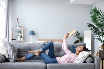 Beautiful smiling woman reading a book and lying on the sofa in the living room.