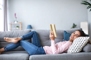 Smiling woman with a book lying on the sofa in the living room.