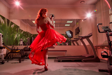 Beautiful girl with makeup, red curly hair and a red dress having training with dumbbells in the gym