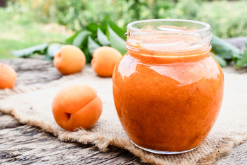A glass jar with homemade apricot jam on a wooden surface near ripe apricot and green leaves.
