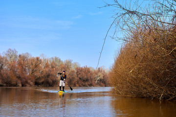 Man on stand up paddle boarding (SUP) paddling along the calm autumn Danube river at sunset against...