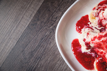 Closeup Vanilla ice cream poured with strawberry or raspberry jam with sliced banana slices on a white plate on a dark wooden table. You can do this at home on vacation