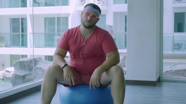Funny fat male in pink glasses and in a pink t-shirt is engaged on a fit ball in the gym depicting a girl. A wonderful man listens to music and dances on the ball in the gym.