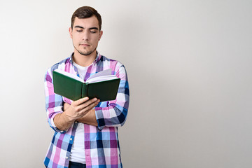 Young man student in plaid shirt reading book on grey background with copy space