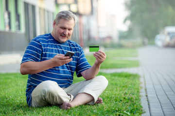 A satisfied man makes online purchases using a credit card and a smartphone, sitting on the lawn in the city with a laptop.