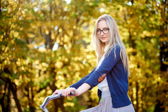 Portrait of blond long-haired attractive tourist woman in glasses, skirt and blouse posing on modern lady bicycle on lit by autumn sun park on bright colorful golden bokeh trees background.