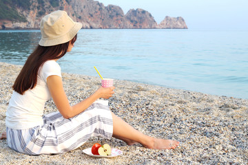 Girl sitting on the beach holds a paper cup and a plate of apples.