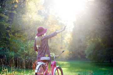 Back view of slim blond fashionable long-haired attractive girl in short dress and pink hat with raised arm at lady bicycle on paved summer park alley, enjoying beautiful evening sunset