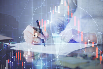 Hands writing information about stock market in notepad. Forex chart holograms in front. Concept of research. Multi exposure