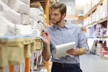 Young man shopping or working in a hardware warehouse standing checking supplies on his tablet.