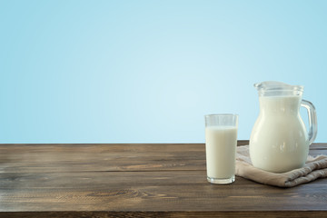 Glass of fresh milk and jug on wooden tabletop with blue wall as background.