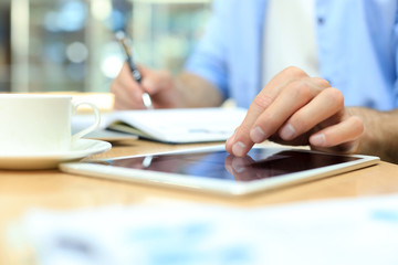 Man writing work plan with pen on paper notebook, browsing internet on digital tablet on office desk.