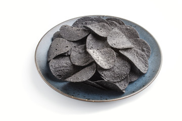 Black potato chips with charcoal on a blue ceramic plate isolated on white background. side view.