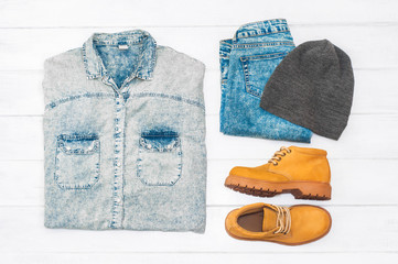 Set of clothes on white wooden background. Denim shirt, jeans, cap, and yellow boots. Fashion. Casual style. Warm clothing. Top view. Flat lay