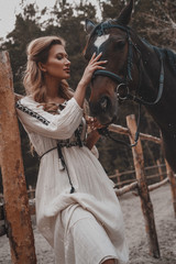 Beautiful and tender young woman wearing the dress is embracing and stroking the horse on the ranch. An attractive rider is posing outdoors near the saddle. Summertime, nature landscape, countryside