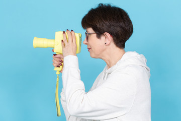 Side view of a middle-aged woman with a short haircut in a sweater and glasses holding a yellow vintage movie camera on a blue background. Concept of video shooting