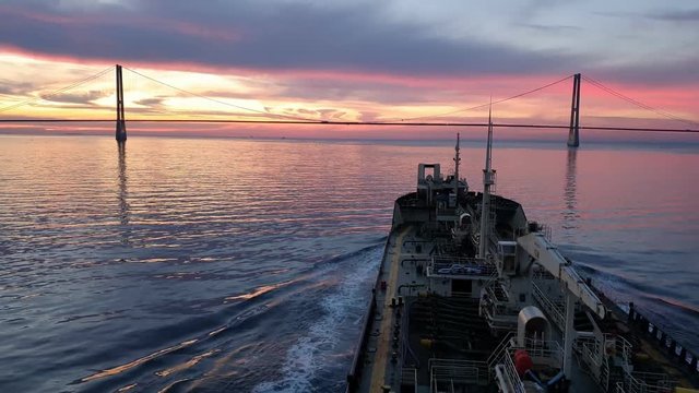 modern bulk carrier sails to huge bridge silhouette under purple and orange sunset sky reflected in deep blue river