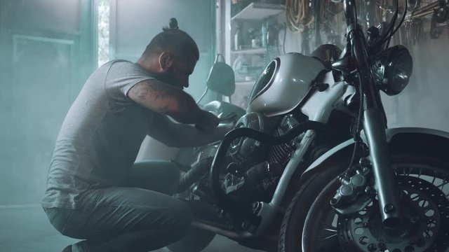 biker with a motorcycle in an atmospheric garage. a bright-looking man with a beard and tattoos is preparing his iron horse for the ride.