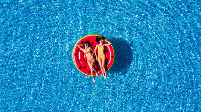 Aerial view of two women lying on inflatable watermelon mattrass floating and relaxing in swimming pool