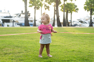 A little girl of 1 year old running on a green meadow in a park among palm trees and cutting a red ribbon