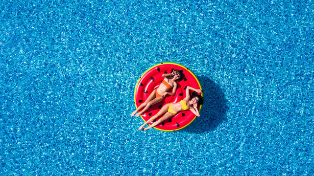 Aerial view of two women lying on inflatable watermelon mattrass floating and relaxing in swimming pool