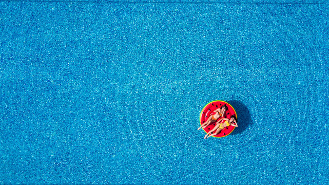 Aerial view of two women lying on inflatable watermelon mattrass floating and relaxing in swimming pool